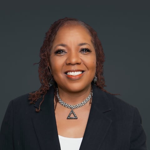 Professional headshot of a smiling woman with curly reddish-brown hair wearing a black blazer and decorative triangle pendant necklace