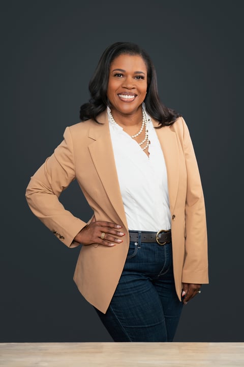 Professional portrait of a woman in a tan blazer and white top smiling against a gray background