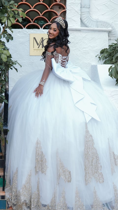 Woman wearing white ball gown with beaded detailing, tiara, and sparkly jewelry poses against white wall with decorative trellis