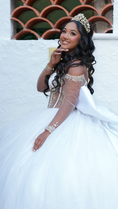 Woman in white wedding dress with embellished bodice, tiara, and long black wavy hair smiling at camera against decorative brick background