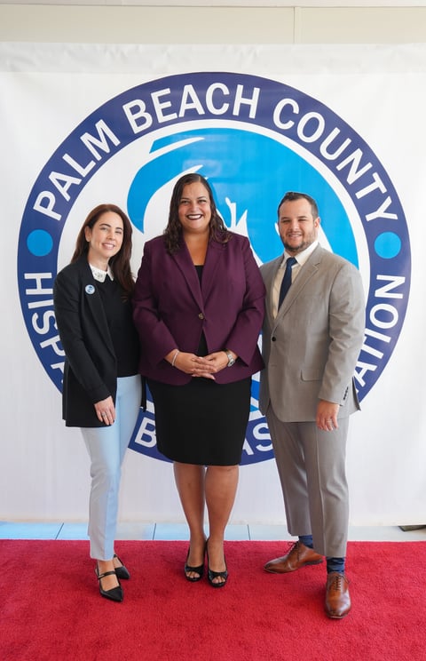 Three professional people stand on red carpet in front of Palm Beach County Tourism logo backdrop