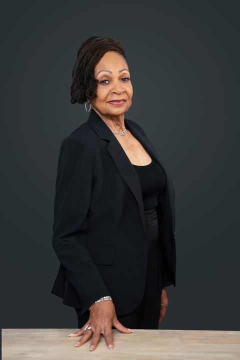 Professional headshot of a woman wearing a black blazer and black top, posing against a gray background with her hand resting on a wooden table