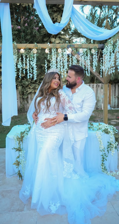 Couple in white wedding attire posing at decorated outdoor venue with blue draped fabric and hanging floral arrangements