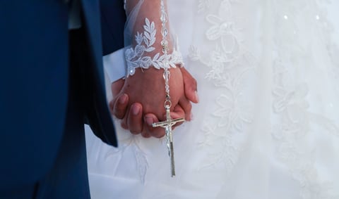 Bride and groom holding hands with decorative rosary, showing wedding rings during ceremony