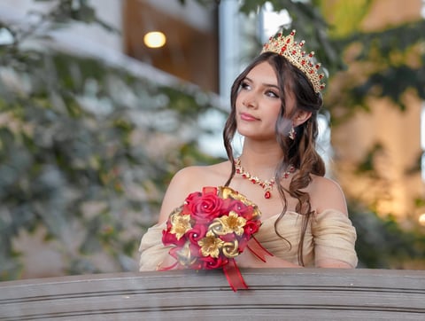 Woman wearing a tiara and off-shoulder dress holds a pink and yellow flower bouquet at an elegant indoor venue