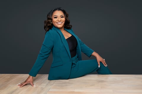 Woman in teal business suit posing confidently while sitting on a wooden table against a gray background