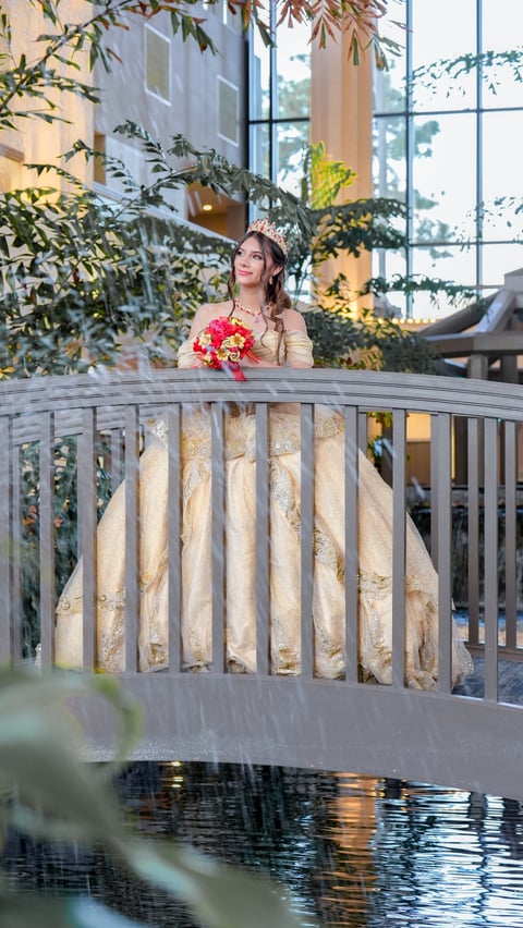 A bride in a beige wedding dress and floral crown holds a red bouquet while standing on a wooden bridge near plants and water