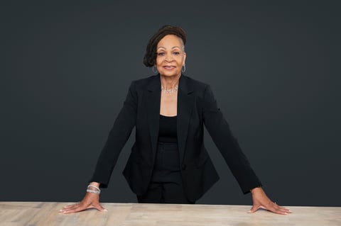 Professional woman in black blazer leaning on desk against gray background