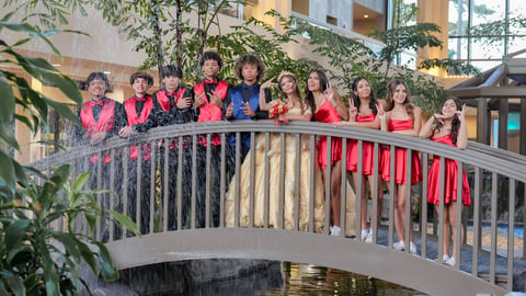Group of young women in red and pink outfits standing together on a curved indoor bridge with railings and green plants