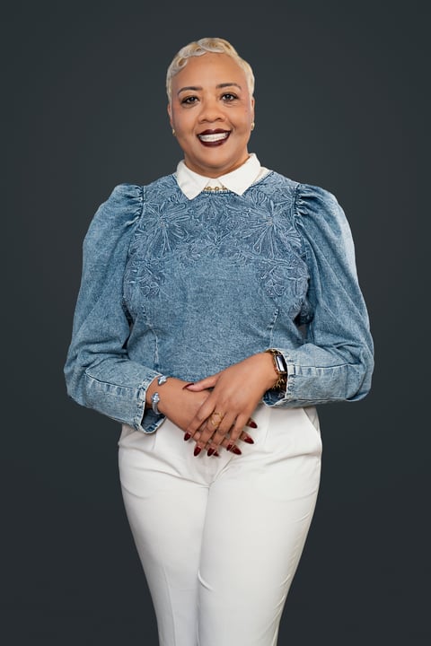 Portrait of a woman with short blonde hair smiling, wearing a blue denim top with puffed sleeves and white pants against a dark gray background