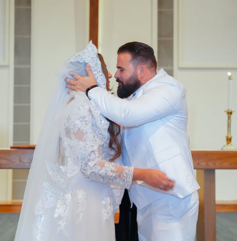 A man in a light blue shirt embraces a bride in a white lace gown with veil in a church sanctuary