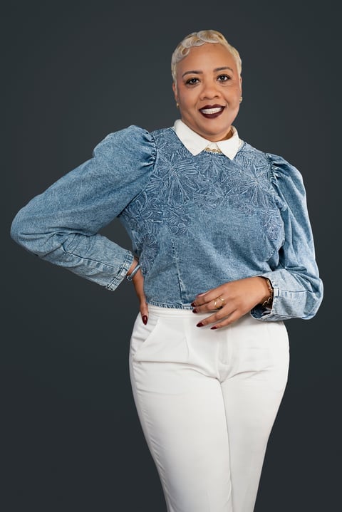 Woman with short blonde hair wearing a blue denim jacket with puffed sleeves, white collar, and white pants, posing against a dark gray background