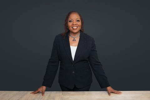 Professional woman in black blazer leaning on desk against gray background