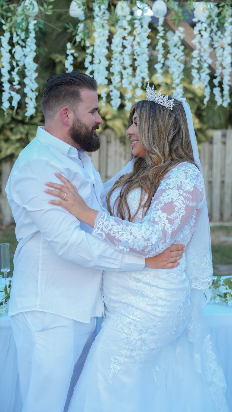 Bride and groom in white formal attire embrace under hanging floral garland at wedding ceremony