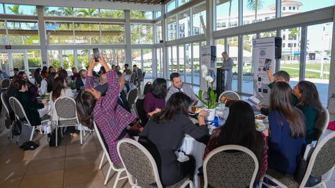A group of people seated at tables during an indoor event in a bright glass-walled venue with outdoor views