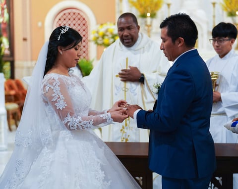 Bride in white lace gown and veil exchanges rings with groom in blue suit during church wedding ceremony with priest and groomsmen present