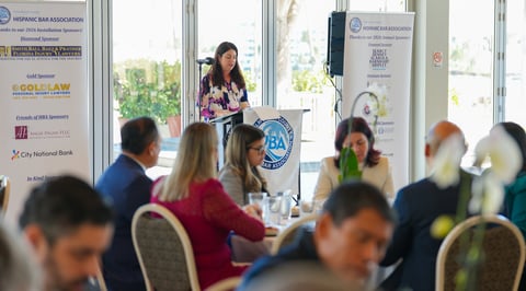 Woman presenting at a professional conference or business seminar with attendees seated at tables and sponsor logos on display