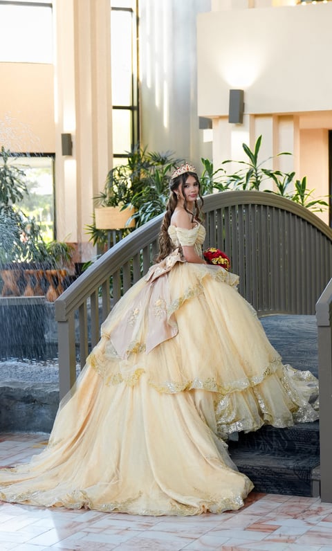 Woman in cream-colored ball gown with tiara standing on curved bridge in modern interior with plants and windows