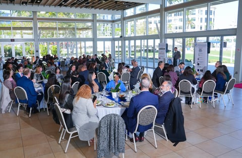 Large group of people seated at round tables in a bright, modern venue with floor-to-ceiling windows overlooking green spaces