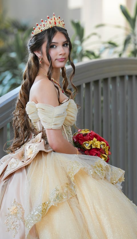 Woman wearing a cream ball gown with gold tiara and red bouquet, standing on a balcony with plants in background