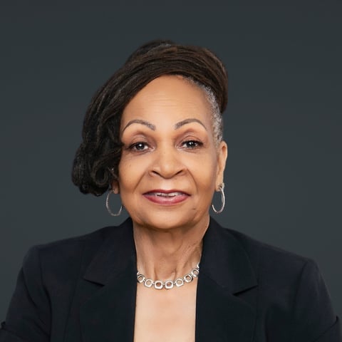 Professional headshot of a woman with braided hair, wearing a black blazer and decorative necklace against a gray background