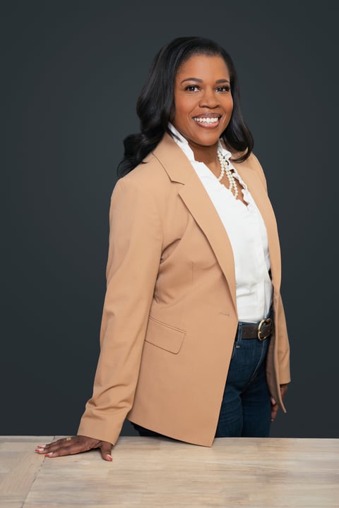 Professional woman smiling, wearing a beige blazer and white top, posing at a wooden table against a gray background