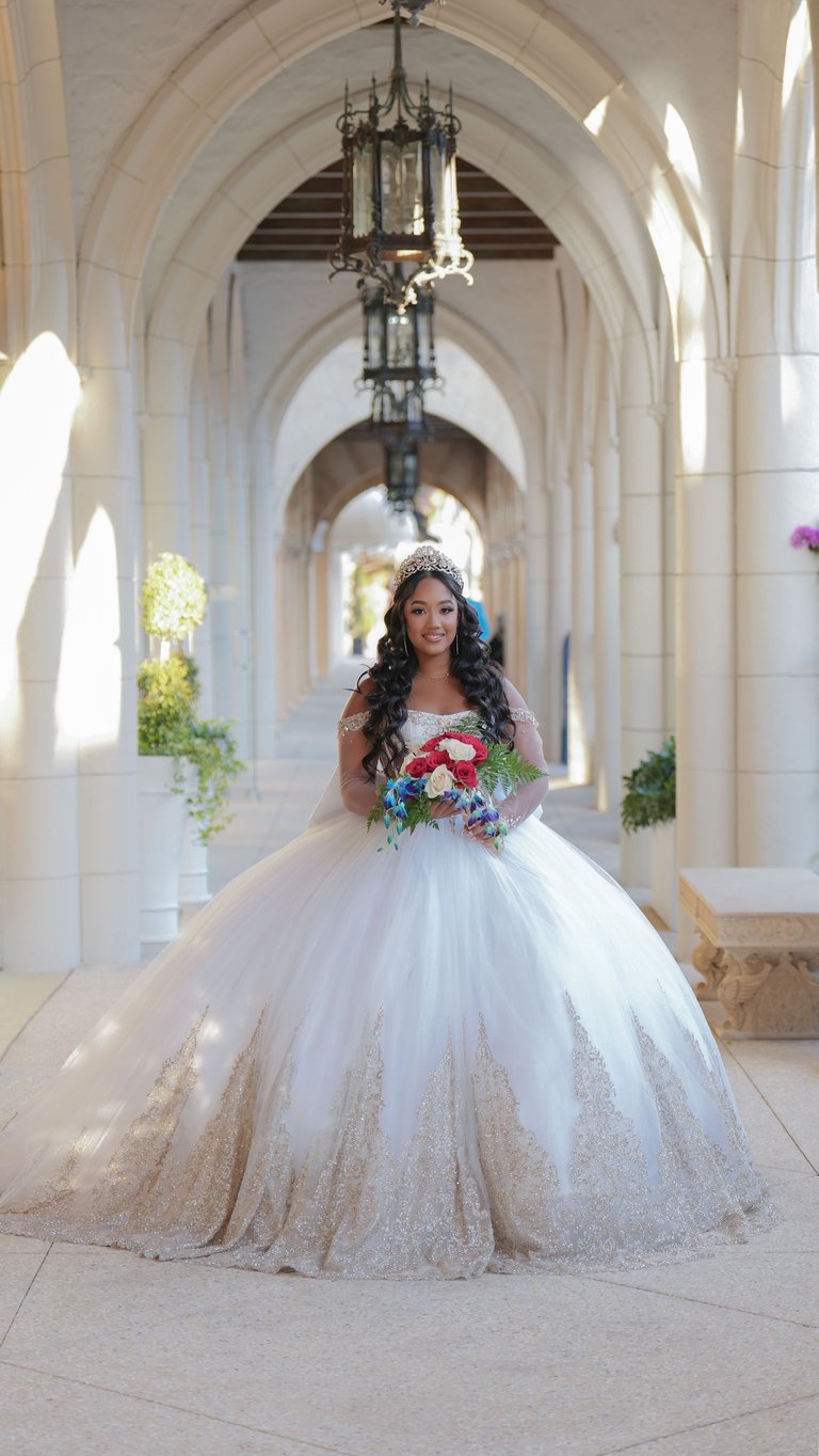Bride in white ball gown with colorful bouquet standing in arched hallway with ornate chandelier