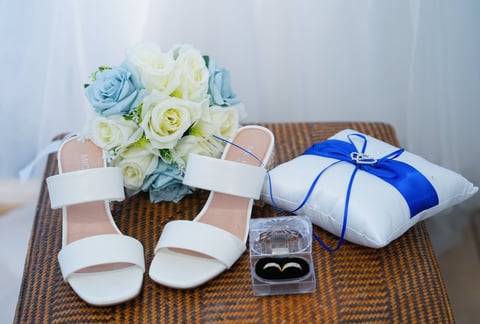 Bridal accessories arranged on a table with white sandals, blue and white gift box, wedding rings in a box, and blue and white floral bouquet