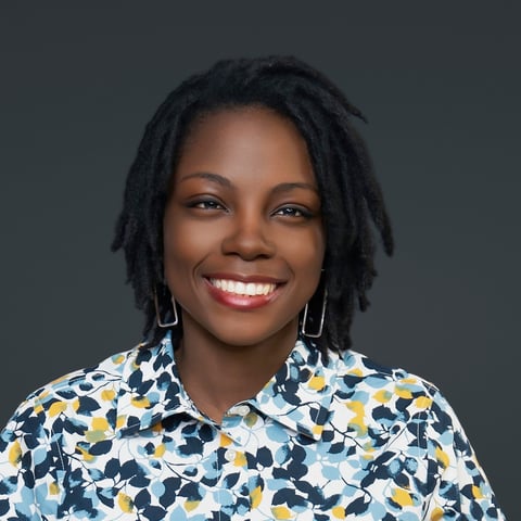 Portrait of a smiling woman with dreadlocks wearing a blue and yellow floral patterned shirt against a gray background