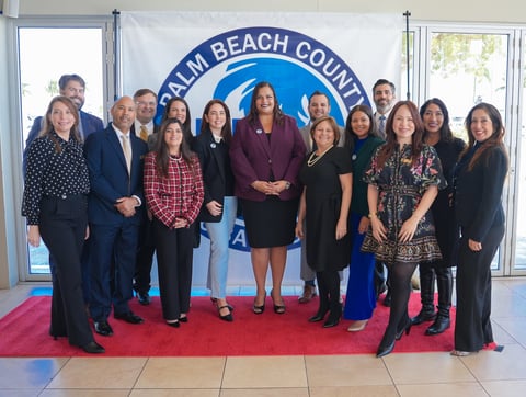 Group of professional women and men standing on a red carpet in front of a Palm Beach County logo banner
