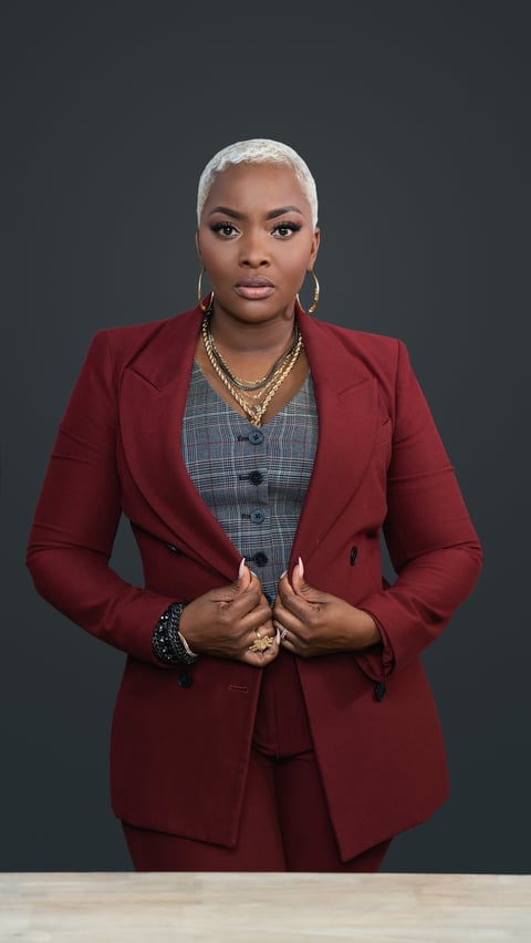Professional woman wearing a burgundy blazer and plaid vest with white short hair and gold chain necklace against dark background