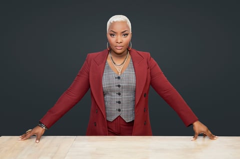 Professional woman with white hair wearing a burgundy blazer and gray vest leaning on a wooden table against a dark gray background
