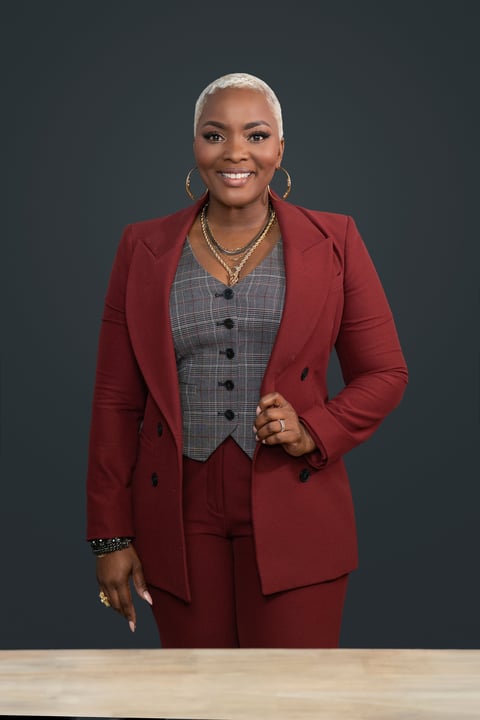 Professional portrait of a woman in a burgundy suit with a gray plaid vest, gold necklace, and hoop earrings, smiling at the camera against a dark background