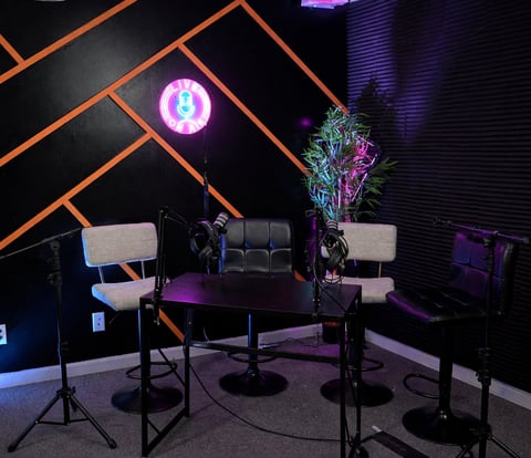 Modern podcast studio with neon orange geometric wall, two gray chairs, black desk, purple lighting, microphones, and illuminated pink logo above