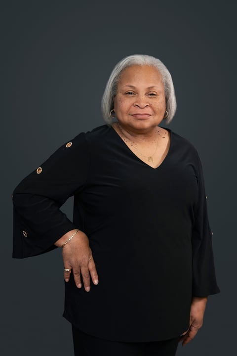 Portrait of a woman with white hair wearing a black v-neck top against a gray background, smiling at the camera