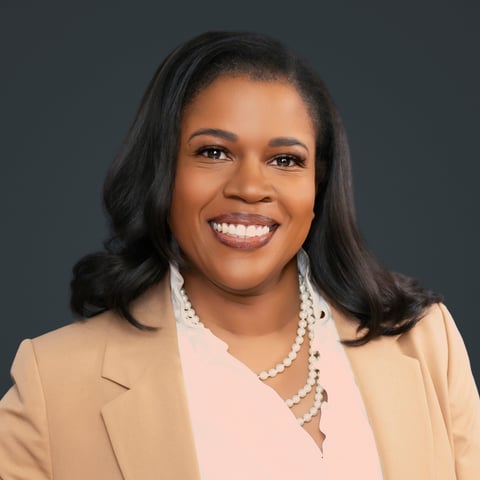 Professional headshot of a smiling woman with long dark hair wearing a beige blazer and pearl necklace against a gray background