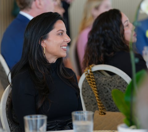Woman with long dark hair smiling at an indoor event, wearing black top with glasses of water on table in foreground