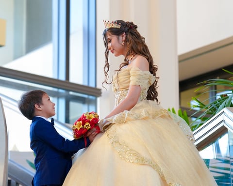 Young boy in navy suit holding red and yellow flowers next to girl in yellow ball gown with off-shoulder sleeves in bright indoor venue