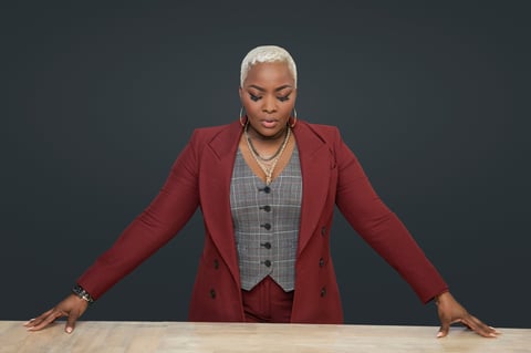 Woman with white hair wearing red blazer and gray vest leaning on table against dark background