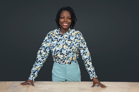 Smiling woman with long dark hair wearing a blue floral blouse and light blue pants, leaning on a wooden desk against a dark background