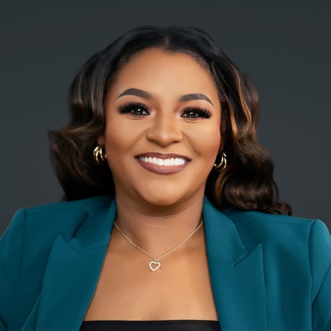 Professional headshot of a woman smiling at the camera, wearing a teal blazer and heart pendant necklace against a gray background