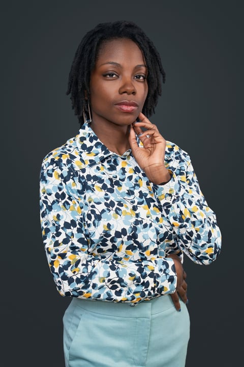 Woman with black braided hair wearing a patterned white, navy, and yellow shirt with light blue pants, hand raised to chin