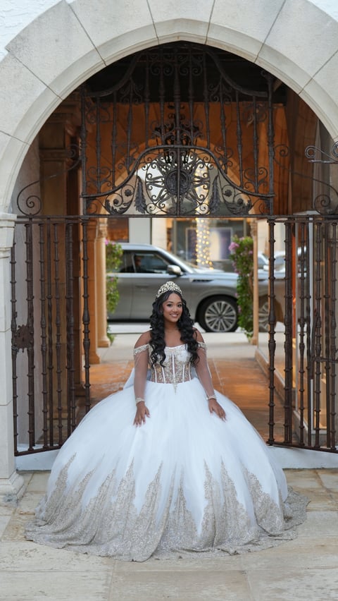 Woman in white ball gown with tiara standing under ornate stone archway with iron gates