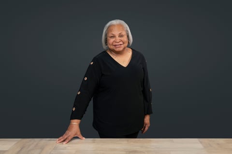 Smiling woman with white hair wearing a black v-neck top, standing at a wooden table against a gray background