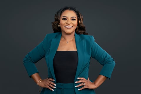 Professional woman in teal blazer and black top smiling with hands on hips against gray background