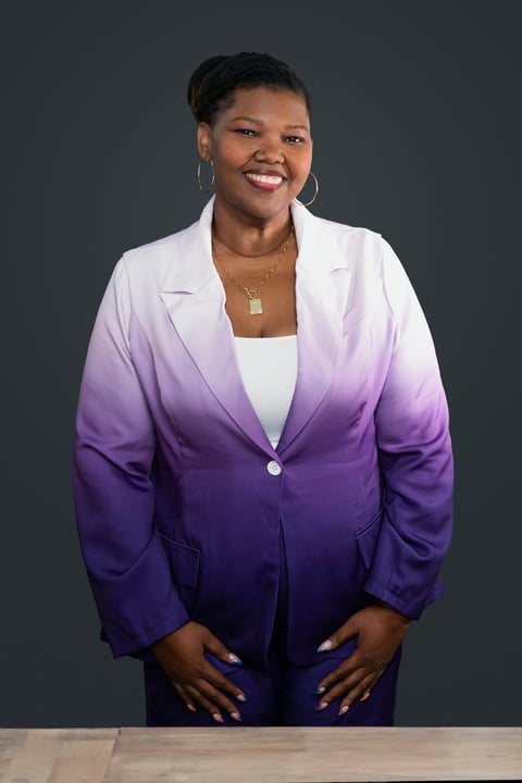 Professional headshot of a woman wearing an ombre purple and white blazer with a white undershirt, smiling at the camera against a gray background